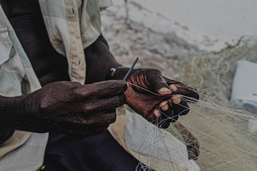 A person is skillfully working with a fishing net, holding a tool in their hands. The focus is on the hands, which are dark and contrasted against the lighter background.