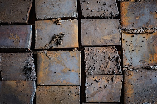 Rusty metal plates arranged in a grid pattern, with a rough texture and corrosion visible on the surface. The plates exhibit signs of wear and oxidation, giving a weathered appearance.