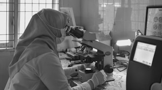 A person wearing a hijab and gloves is examining a sample under a microscope in a laboratory setting. The room is equipped with laboratory instruments and a window with bars lets in natural light. A monitor displays some data in the foreground.