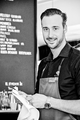 A man in a restaurant setting wears a dark shirt and an apron. He appears to be a waiter or staff member, holding a white cloth and a notepad with a pen. The background shows part of a menu board with blurred text.