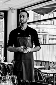 A person standing in a restaurant setting wearing a dark apron and holding a small notepad, with tables set with glassware in the foreground and large windows in the background.