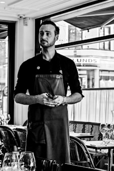 A person standing in a restaurant setting wearing a dark apron and holding a small notepad, with tables set with glassware in the foreground and large windows in the background.