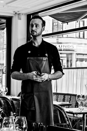 A person standing in a restaurant setting wearing a dark apron and holding a small notepad, with tables set with glassware in the foreground and large windows in the background.