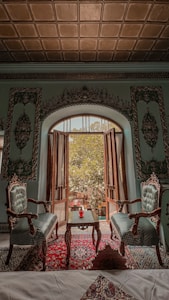 An ornate room featuring two upholstered chairs and a wooden table set with a red vase. The room has an arched wooden door leading to a sunlit garden view. The walls and ceiling are decorated with intricate patterns, and a colorful carpet covers the floor.