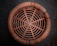 A circular woven rattan stool is seen from above, showcasing its intricate spiral design. The surrounding ground is tiled with a muted gray-blue color, adding a contrasting background to the warm brown tones of the stool.