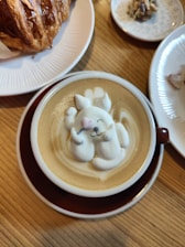 A cup of coffee features intricate latte art depicting a cute animal character with a pink nose and closed eyes, resting on its back. The latte art is surrounded by smooth, creamy coffee. Nearby, a flaky croissant is placed on a white plate, and another small dish contains a bite-sized treat.