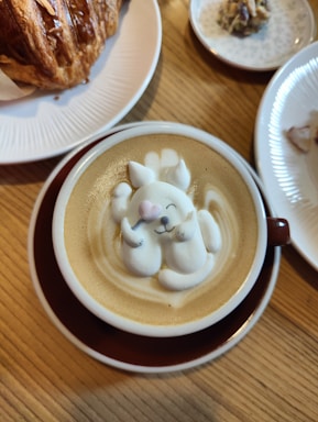 A cup of coffee features intricate latte art depicting a cute animal character with a pink nose and closed eyes, resting on its back. The latte art is surrounded by smooth, creamy coffee. Nearby, a flaky croissant is placed on a white plate, and another small dish contains a bite-sized treat.