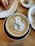 A cup of coffee features intricate latte art depicting a cute animal character with a pink nose and closed eyes, resting on its back. The latte art is surrounded by smooth, creamy coffee. Nearby, a flaky croissant is placed on a white plate, and another small dish contains a bite-sized treat.