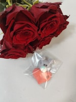 A bouquet of vibrant red roses placed beside a small toy bear holding a red heart, encased in a clear plastic bag.