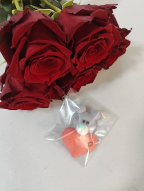A bouquet of vibrant red roses placed beside a small toy bear holding a red heart, encased in a clear plastic bag.