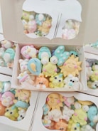 A collection of colorful, cute cartoon-like characters in pastel shades, packed inside cardboard boxes with clear lids. The characters have cheerful, smiling faces and are shaped like various animals and objects.