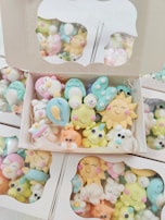 A collection of colorful, cute cartoon-like characters in pastel shades, packed inside cardboard boxes with clear lids. The characters have cheerful, smiling faces and are shaped like various animals and objects.