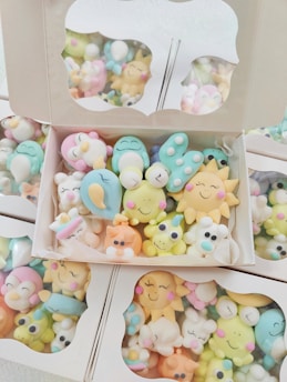 A collection of colorful, cute cartoon-like characters in pastel shades, packed inside cardboard boxes with clear lids. The characters have cheerful, smiling faces and are shaped like various animals and objects.