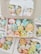 A collection of colorful, cute cartoon-like characters in pastel shades, packed inside cardboard boxes with clear lids. The characters have cheerful, smiling faces and are shaped like various animals and objects.