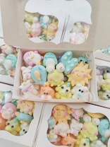 A collection of colorful, cute cartoon-like characters in pastel shades, packed inside cardboard boxes with clear lids. The characters have cheerful, smiling faces and are shaped like various animals and objects.