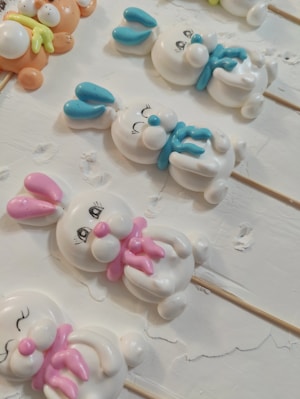 Cute decorative figures resembling bunnies are arranged on a flat surface. These figures are crafted with white material, featuring colorful accents on their ears and scarves, such as pink, blue, and orange. The surface appears textured and slightly cracked.