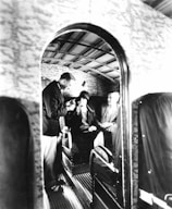 A vintage airplane interior with three people seated, two men and one woman. The man on the left, dressed in a dark suit, is standing and possibly offering refreshments or assistance. The woman in the center is wearing a hat, and the man on the right is seated comfortably. The walls and ceiling have a textured design, and the overall atmosphere gives a sense of early air travel luxury.