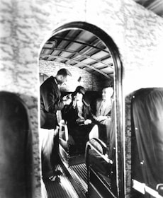 A vintage airplane interior with three people seated, two men and one woman. The man on the left, dressed in a dark suit, is standing and possibly offering refreshments or assistance. The woman in the center is wearing a hat, and the man on the right is seated comfortably. The walls and ceiling have a textured design, and the overall atmosphere gives a sense of early air travel luxury.