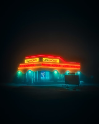 A neon-lit store stands illuminated in the night. The bright lights, in contrasting red and yellow hues, advertise services like payday advances and loans. The scene is enveloped in darkness, creating a mysterious and moody atmosphere, with fog adding to the surreal ambiance.