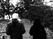 Two people walk down a leaf-covered path in a wooded area, framed by dense trees. The scene appears serene and contemplative, captured in black and white, which enhances the contrast between the figures and the natural background.