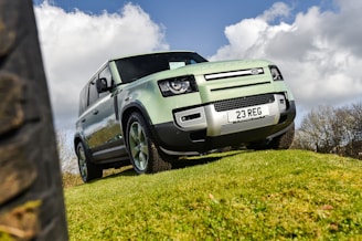 A green 4x4 vehicle is parked on a grassy hill against a backdrop of a partly cloudy sky. The vehicle has a robust and rugged design, featuring a license plate reading '23 REG'. The angle of the shot emphasizes the vehicle’s size and off-road capabilities.