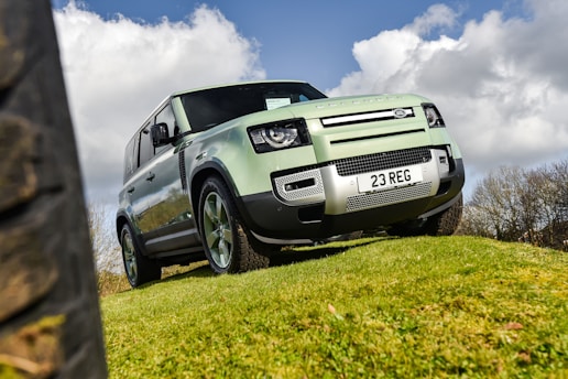A green 4x4 vehicle is parked on a grassy hill against a backdrop of a partly cloudy sky. The vehicle has a robust and rugged design, featuring a license plate reading '23 REG'. The angle of the shot emphasizes the vehicle’s size and off-road capabilities.