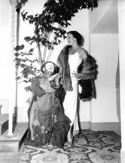 A person wearing a patchwork outfit and clown makeup is interacting with a well-dressed woman in an elegant gown and fur stole. They are indoors, with a decorative plant in the background and an intricately patterned carpet underfoot.