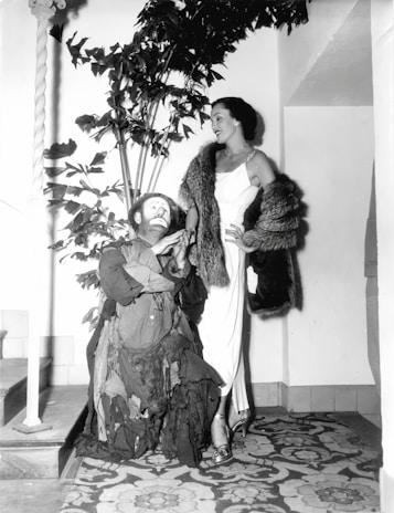 A person wearing a patchwork outfit and clown makeup is interacting with a well-dressed woman in an elegant gown and fur stole. They are indoors, with a decorative plant in the background and an intricately patterned carpet underfoot.