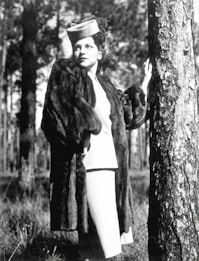 A woman in a vintage outfit stands in a forest, wearing a fur coat and a stylish hat. She leans against a tree, looking into the distance with a contemplative expression. The black and white image highlights the textures of her clothing and the tree bark against a backdrop of tall trees.