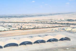 Aerial view of a desert landscape with a small urban area surrounded by arid land. Five aircraft hangars with planes are visible in the foreground, with rows of houses and some green patches in the background extending towards a vast expanse of desert.