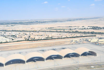 Aerial view of a desert landscape with a small urban area surrounded by arid land. Five aircraft hangars with planes are visible in the foreground, with rows of houses and some green patches in the background extending towards a vast expanse of desert.