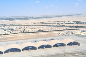 Aerial view of a desert landscape with a small urban area surrounded by arid land. Five aircraft hangars with planes are visible in the foreground, with rows of houses and some green patches in the background extending towards a vast expanse of desert.