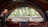 A room with a large, decorative window shaped like a fan that provides a view of a lush green outdoor area with tall plants. The interior features a cozy setting with colorful cushions and fabric-covered tables, along with a hanging red lamp.