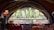 A room with a large, decorative window shaped like a fan that provides a view of a lush green outdoor area with tall plants. The interior features a cozy setting with colorful cushions and fabric-covered tables, along with a hanging red lamp.
