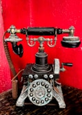 A vintage rotary telephone with intricate metal detailing set against a vibrant red background. The phone features a classic dial with numbers and symbols, ornate design elements, and a traditional receiver resting on top.
