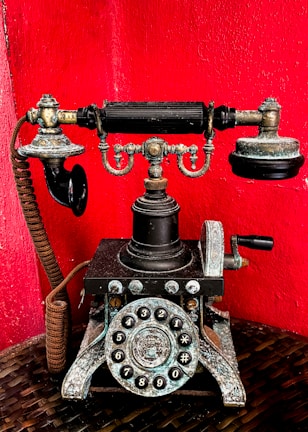 A vintage rotary telephone with intricate metal detailing set against a vibrant red background. The phone features a classic dial with numbers and symbols, ornate design elements, and a traditional receiver resting on top.