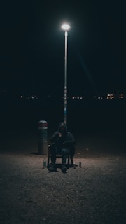 A person is sitting alone on a chair under a bright streetlamp in a dark and empty area. The scene is mostly obscured by darkness, highlighting the isolation of the individual. A few bottles are placed on the ground nearby, adding to the solitary mood.
