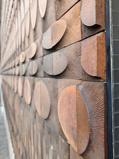 A close-up view of a textured wooden wall featuring numerous semicircular wooden pieces arranged in a staggered pattern. The surface of the wooden pieces highlights the grain and rough texture, creating a tactile and visually intriguing effect.