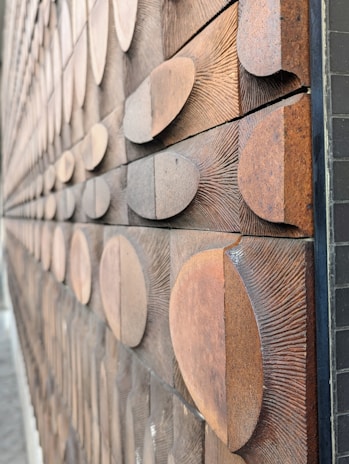 A close-up view of a textured wooden wall featuring numerous semicircular wooden pieces arranged in a staggered pattern. The surface of the wooden pieces highlights the grain and rough texture, creating a tactile and visually intriguing effect.