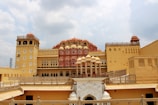 A grand palace structure featuring intricate architecture with multiple levels, rich embellishments, and a distinctive pink and yellow facade. The palace has numerous small windows arranged in a grid-like pattern, adorned with decorative elements typical of Rajasthani architecture. The sky above is partly cloudy, contributing to a dramatic backdrop.