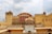 A grand palace structure featuring intricate architecture with multiple levels, rich embellishments, and a distinctive pink and yellow facade. The palace has numerous small windows arranged in a grid-like pattern, adorned with decorative elements typical of Rajasthani architecture. The sky above is partly cloudy, contributing to a dramatic backdrop.