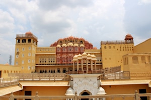 A grand palace structure featuring intricate architecture with multiple levels, rich embellishments, and a distinctive pink and yellow facade. The palace has numerous small windows arranged in a grid-like pattern, adorned with decorative elements typical of Rajasthani architecture. The sky above is partly cloudy, contributing to a dramatic backdrop.