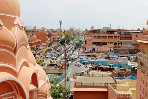 A bustling city scene featuring pink-hued architecture characteristic of Jaipur, India. The image captures a busy street lined with shops and buildings, with a prominent Indian flag. Various vehicles, including cars and scooters, navigate through the traffic, while pedestrians are visible on the sidewalks. The setting conveys a lively urban atmosphere.