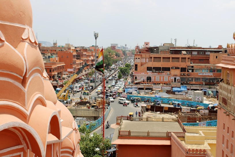 A bustling city scene featuring pink-hued architecture characteristic of Jaipur, India. The image captures a busy street lined with shops and buildings, with a prominent Indian flag. Various vehicles, including cars and scooters, navigate through the traffic, while pedestrians are visible on the sidewalks. The setting conveys a lively urban atmosphere.