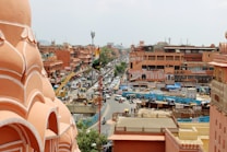 A bustling city scene featuring pink-hued architecture characteristic of Jaipur, India. The image captures a busy street lined with shops and buildings, with a prominent Indian flag. Various vehicles, including cars and scooters, navigate through the traffic, while pedestrians are visible on the sidewalks. The setting conveys a lively urban atmosphere.