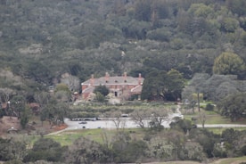 A large estate with a red brick mansion surrounded by dense forest. The building has multiple chimneys and is set in a landscaped garden with a driveway leading to a parking area. The scene is enveloped in lush greenery with trees and bushes in varying shades.