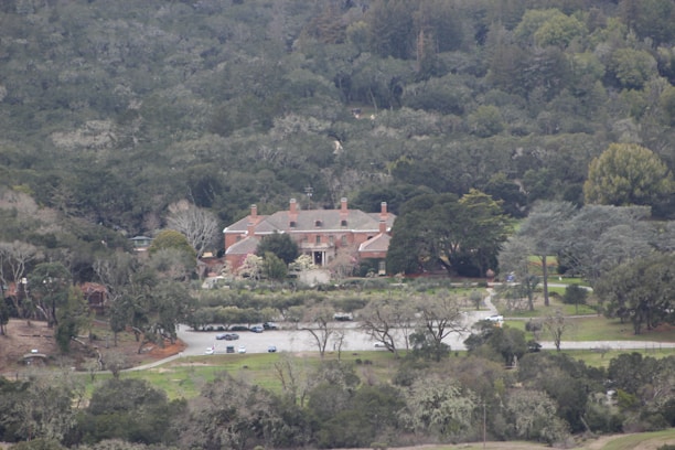 A large estate with a red brick mansion surrounded by dense forest. The building has multiple chimneys and is set in a landscaped garden with a driveway leading to a parking area. The scene is enveloped in lush greenery with trees and bushes in varying shades.