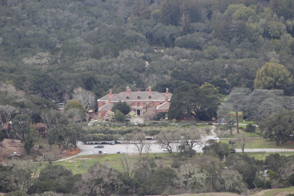 A large estate with a red brick mansion surrounded by dense forest. The building has multiple chimneys and is set in a landscaped garden with a driveway leading to a parking area. The scene is enveloped in lush greenery with trees and bushes in varying shades.