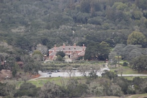 A large estate with a red brick mansion surrounded by dense forest. The building has multiple chimneys and is set in a landscaped garden with a driveway leading to a parking area. The scene is enveloped in lush greenery with trees and bushes in varying shades.