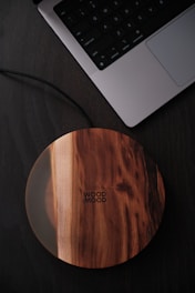A sleek wooden wireless charger with a smooth circular design sits on a dark surface next to an open laptop. The laptop keyboard is partially visible, and the charger features a natural wood grain finish with the words 'Wood Mood' engraved on it.